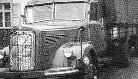 Historische Schwarz-Weiß-Aufnahme eines Lkw mit rundlicher Motorhaube, ein Kind lehnt am vorderen Kotflügel, Gründung des Ferntransportunternehmens 1950
