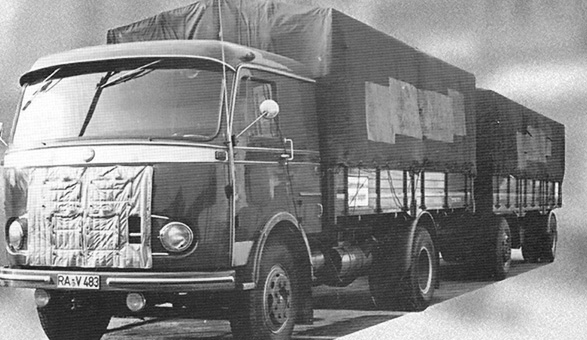 Historische Schwarz-Weiß-Aufnahme eines Lkw mit Anhänger und Planenaufbau, Ausweitung des Unternehmens im Jahr 1955.