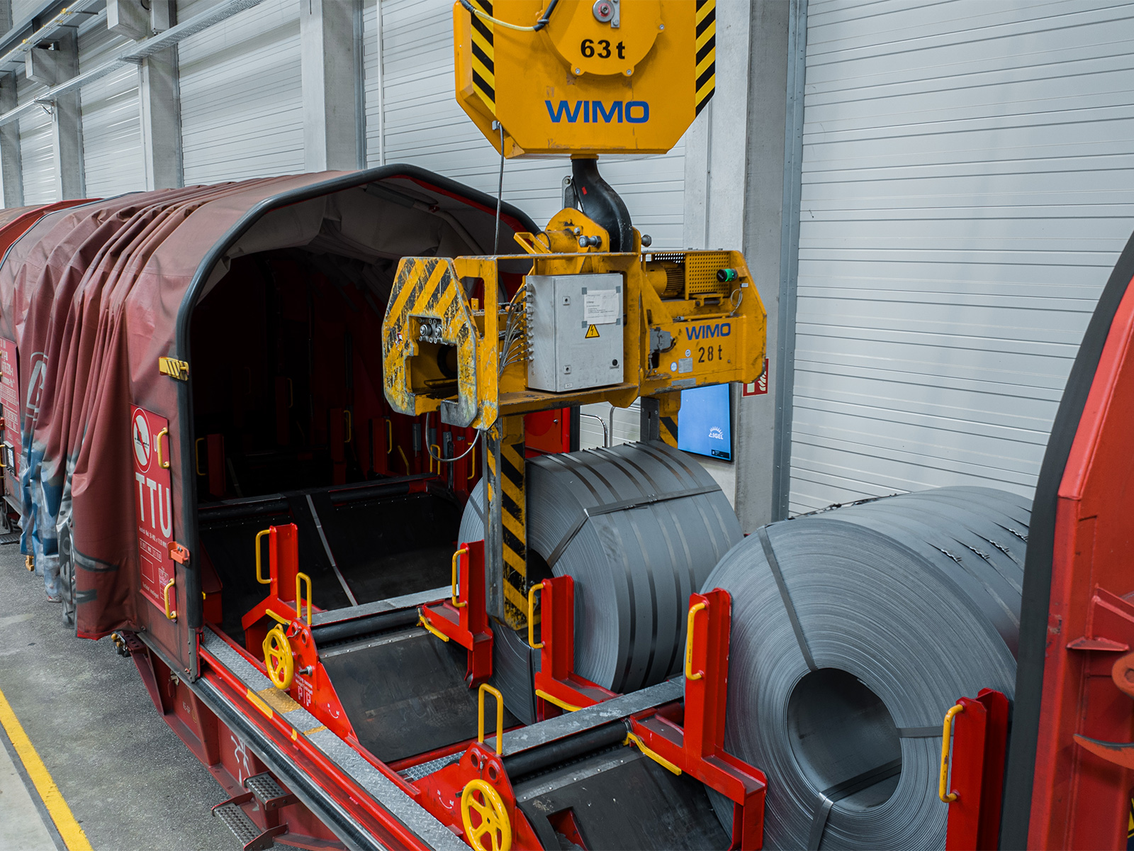 Hallenkran mit spezieller Coilzange beim Verladen schwerer Stahlcoils auf einen Güterwaggon in einer Logistikhalle von Hartmann.