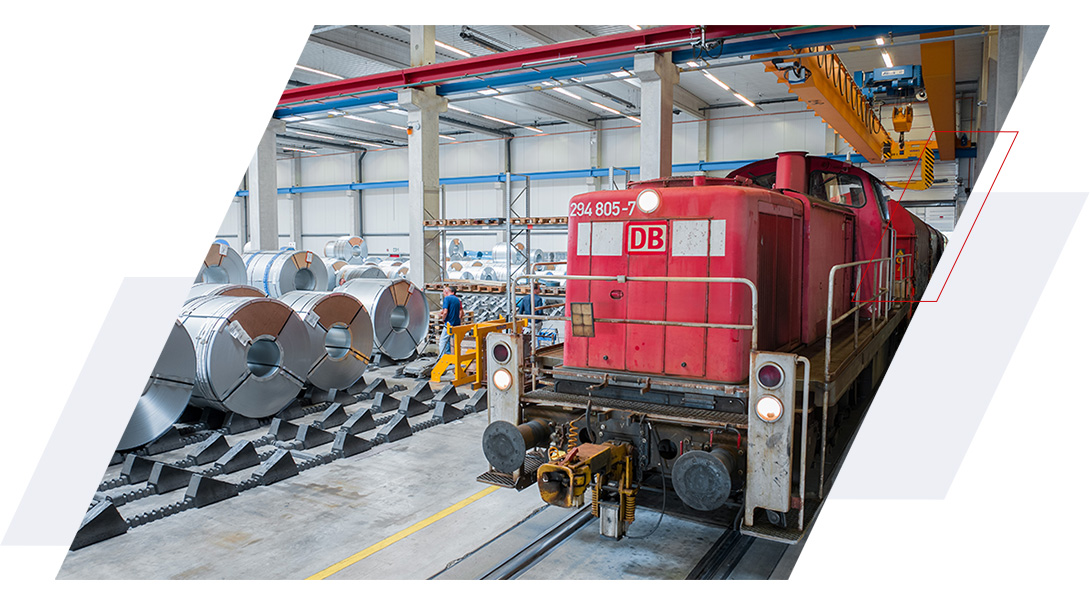 Rote DB-Diesellokomotive auf dem Hallengleis im Coillager von Hartmann, neben gelagerten Stahlcoils und einem Hallenkran.