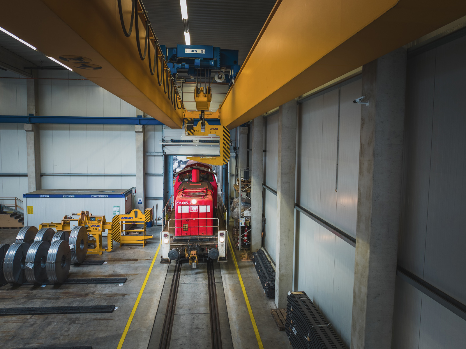 Rote Güterlokomotive in einer Logistikhalle mit Hallenkran und gelagerten Stahlcoils für den Schienentransport.