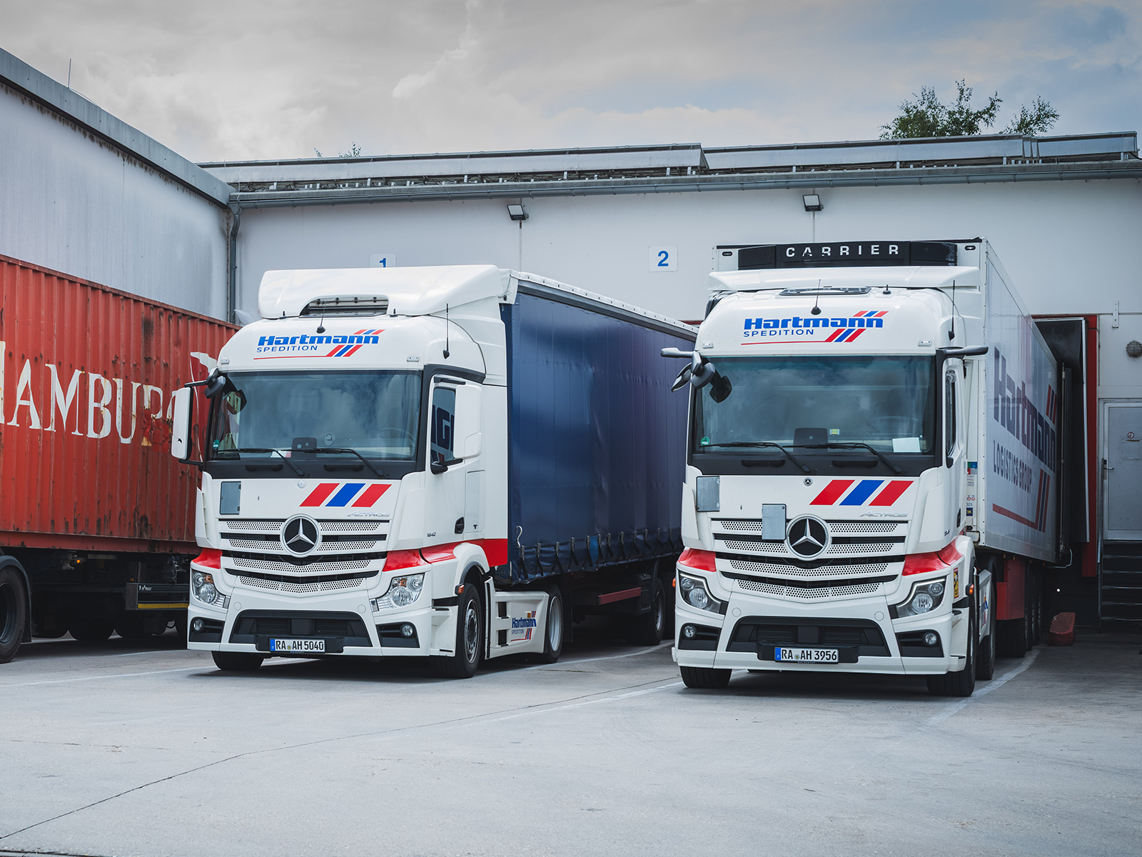 Zwei weiße Hartmann-Lkw vor den Laderampen eines Logistikzentrums als Teil der Transport- und Straßenlogistik.