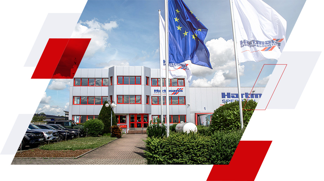 Firmenzentrale der Hartmann Spedition & Logistik mit EU- und Unternehmensflaggen vor dem Gebäude.