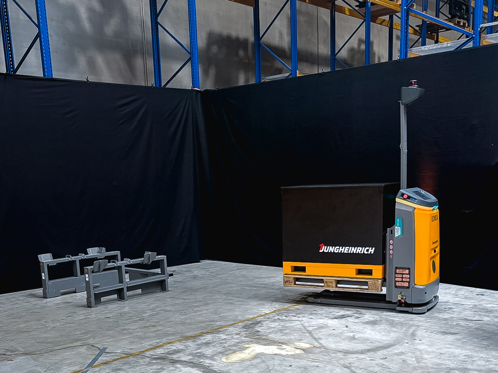Gelb-graues autonomes Flurförderzeug transportiert eine Holzpalette mit einer schwarzen Transportbox in einer Lagerhalle.
