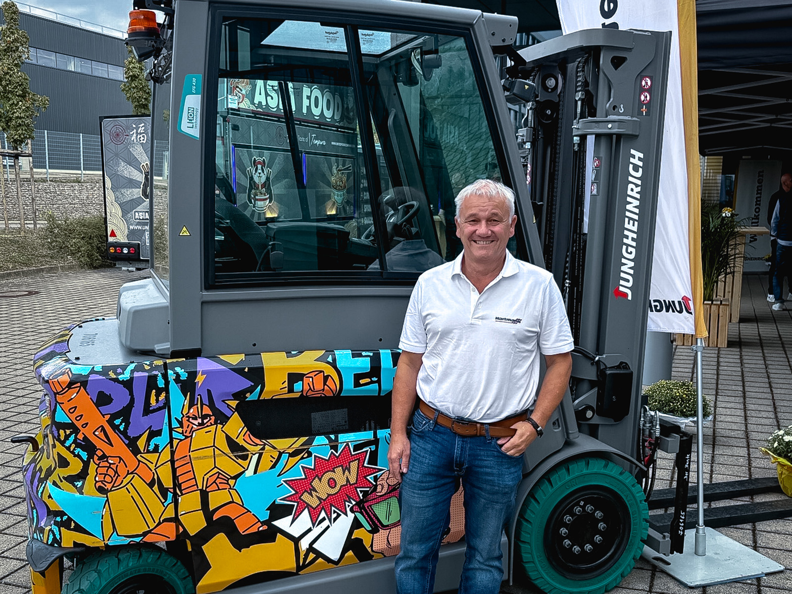 Lächelnder Hartmann Mitarbeiter in weißem Poloshirt steht vor einem Gabelstapler mit buntem Comic-Design auf einem Außengelände.