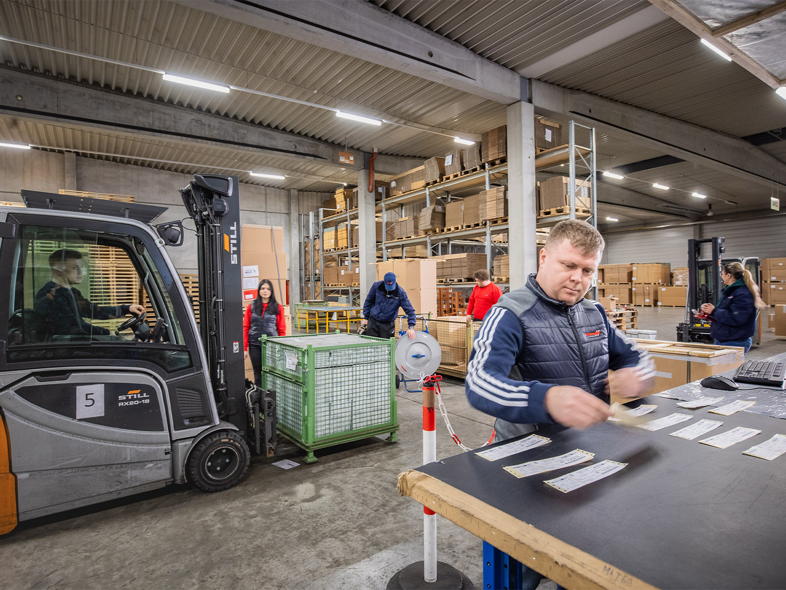 Hartmann-Mitarbeiter bei der Warenkommissionierung und Versandvorbereitung in einer Logistikhalle mit Gabelstaplern und Lagerregalen.
